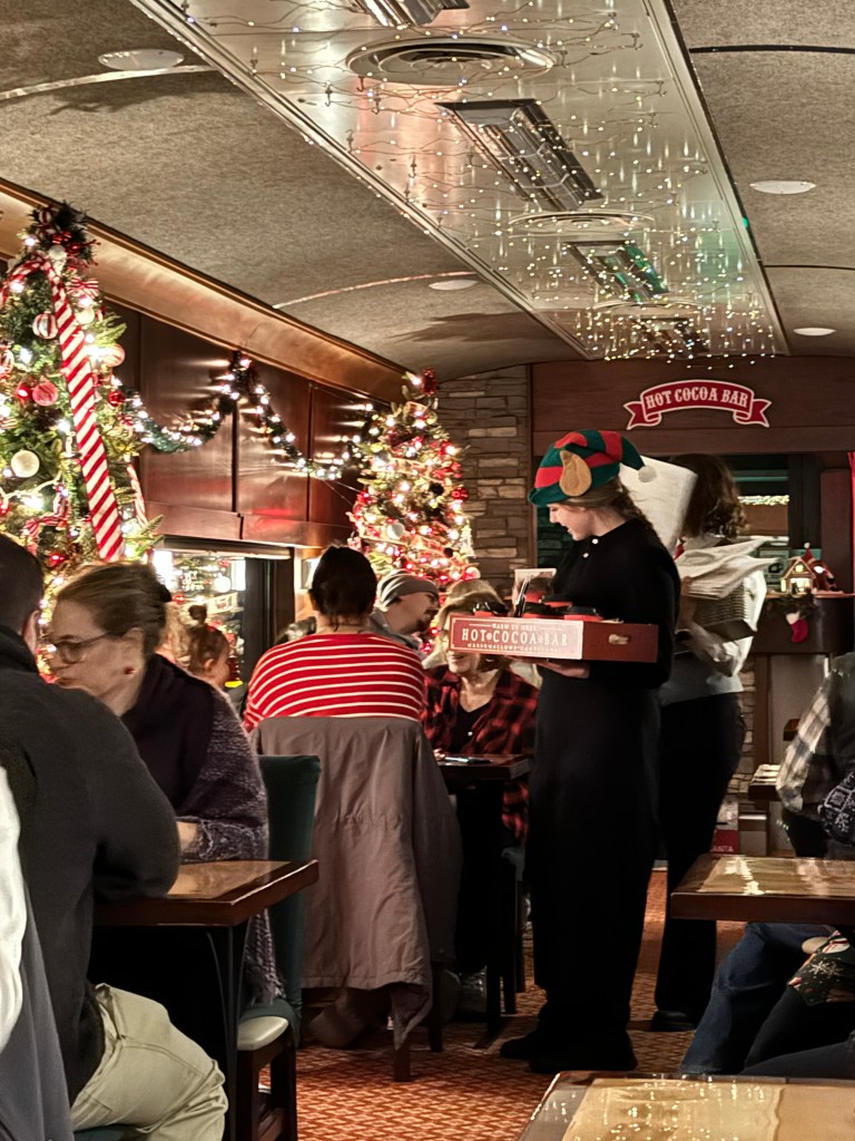 Elves distributing hot cocoa and cookies to passengers aboard Santa's Scenic Railway.